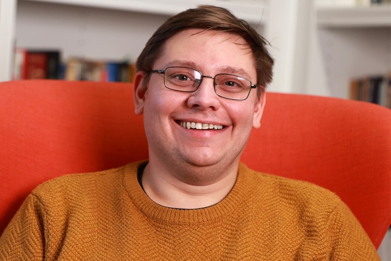 Headshot of James Kneller wearing an orange sweater, sitting in a chair of a different shade of orange.