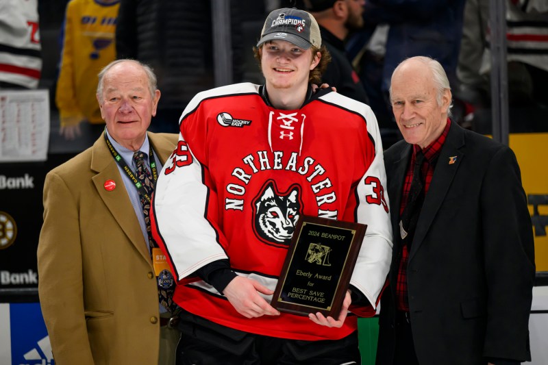 Cameron Whitehead, Northeastern men's hockey goalie, wears his hockey uniform while posing for a picture with two people in suits.