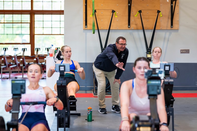 A coach motivates student-athletes while they practice rowing on machines. 