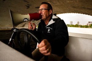 Joe Wilhelm inside of a boat holding the steering wheel with one hand and a megaphone in the other while coaching the team through the megaphone.