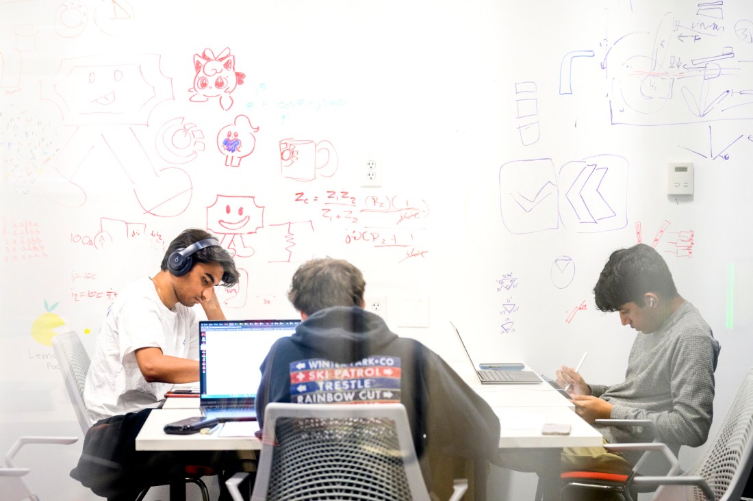 Three students sitting around a table taking notes in front of their laptops. Behind the table is a white board covered in doodles. 