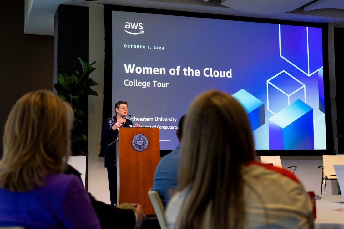 A person speaking at a podium in front of a screen displaying a slide that says 'Women of the Cloud College Tour' with the AWS and Northeastern logos on it.