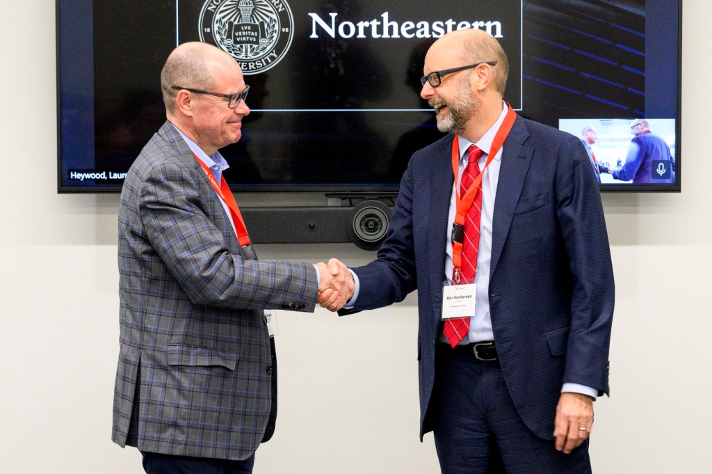 Two men in business attire shake hands in front of a screen displaying the Northeastern University logo.