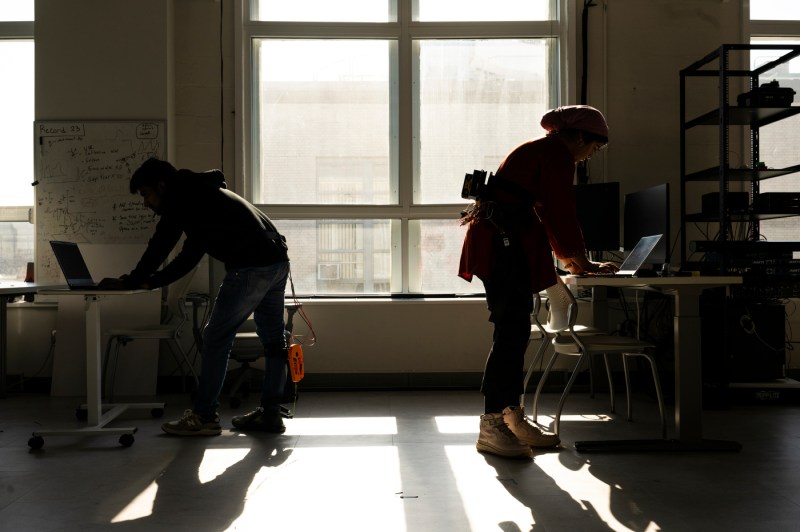 Two people standing with their backs to each other at desks, working on laptops. 