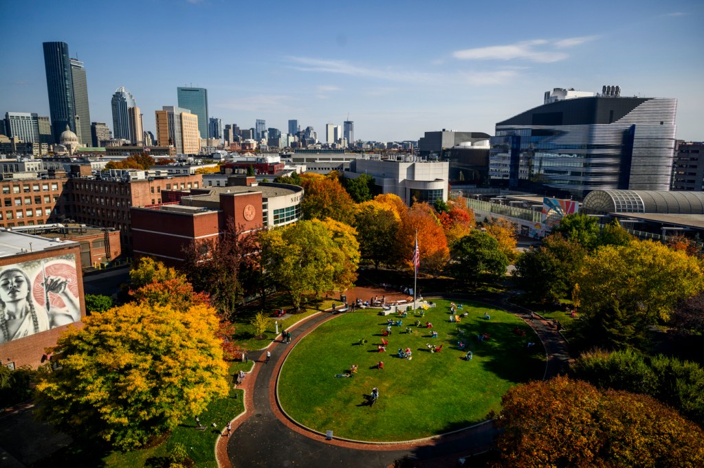 Campus scene with autumn trees, a green lawn, and city buildings in the background.