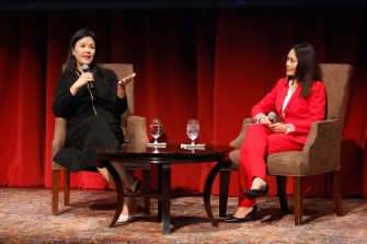 Dr. Rita Ng speaking into a microphone and gesturing with one hand while sitting on a stage in a lecture hall on the Oakland campus.