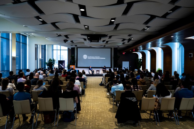 A room full of audience members with a stage and screen at the front for a fireside chat.