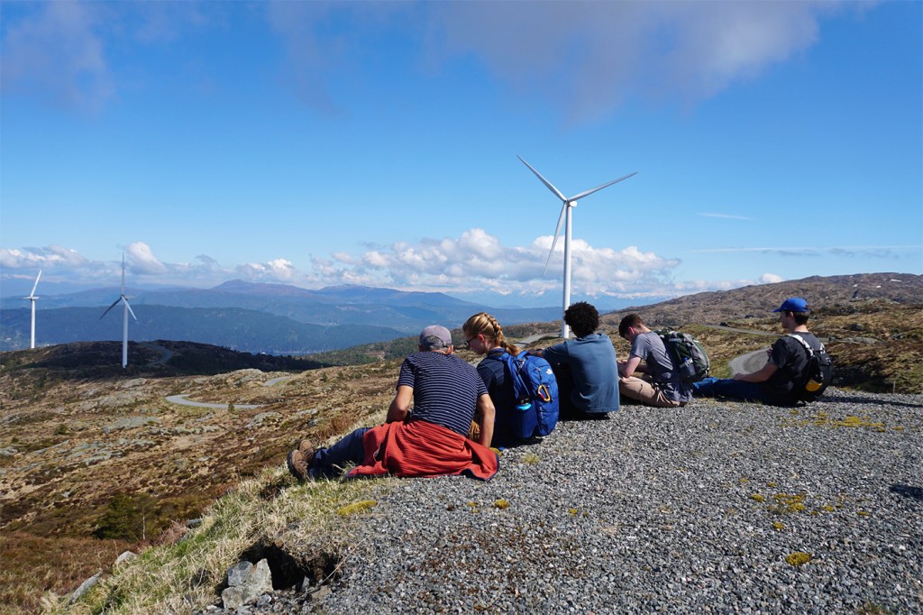 A group of people sitting on the ridge of a hill overlooking several wind turbines.