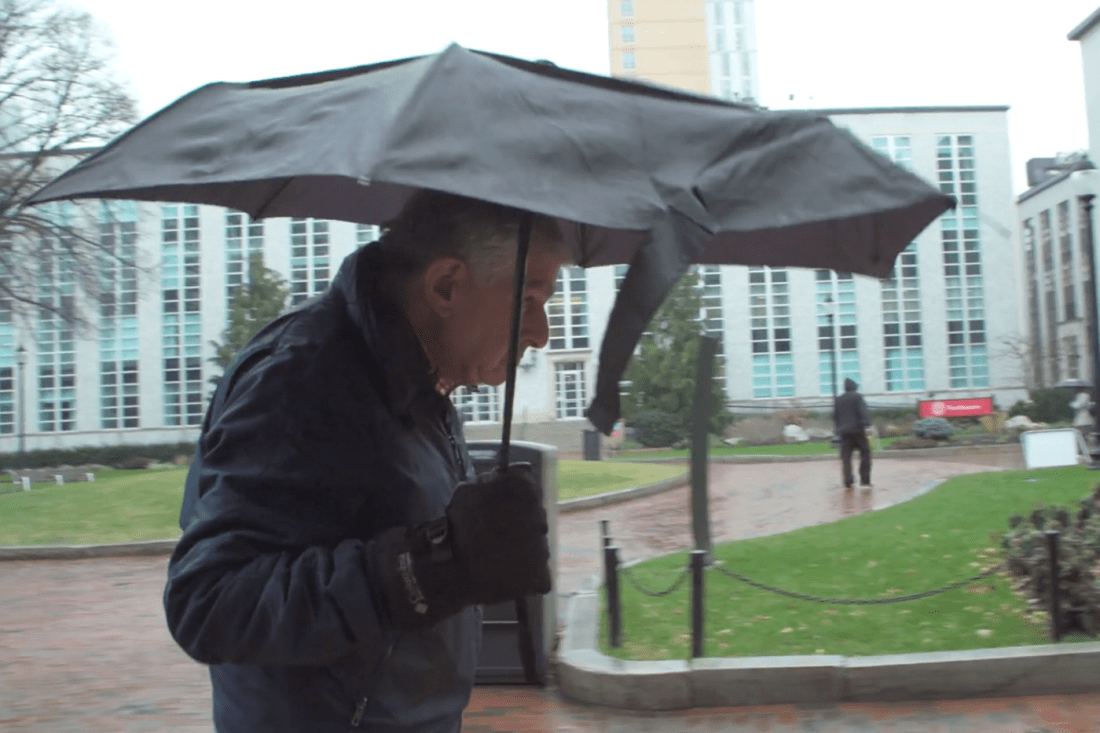 Michael Dukakis walking on Northeastern's Boston campus carrying a black umbrella. 