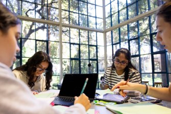 Four people work together at a table inside a library with large windows.