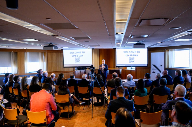 A person speaking at the front of a room full of audience members in front of two screens that say 'Welcome to DMSB Day' on them.