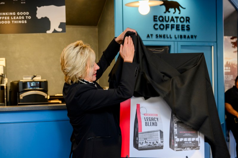 Helen Russell pulling a black curtain off of a poster unveiling the new Northeastern Legacy brand of Equator Coffee.