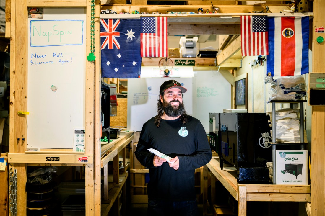 A person stands in a workshop surrounded by flags, tools, and equipment, holding an object and smiling.