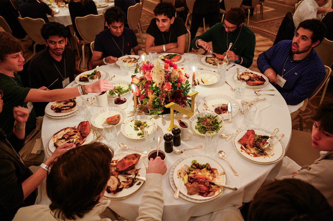 Students seated around a round table enjoying a Thanksgiving meal with floral centerpieces and plates of food.