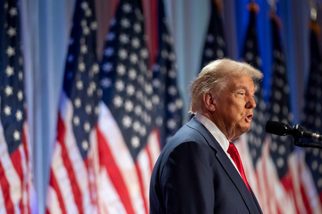 Donald Trump speaking on a stage in front of a row of US flags.