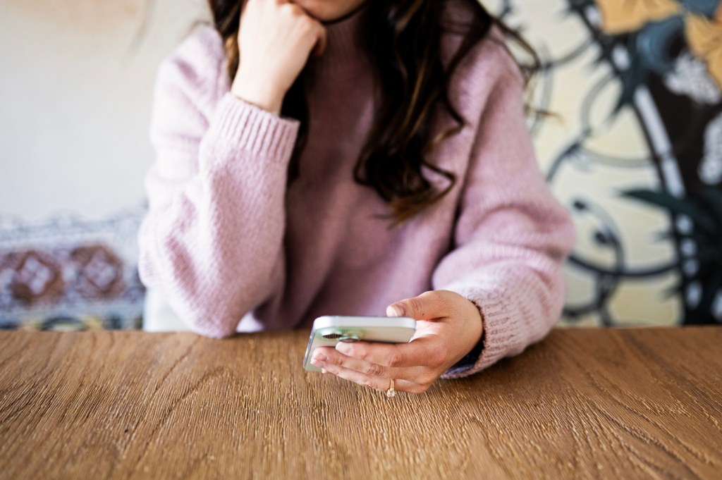 A person wearing a pink sweater sitting at a desk scrolling on their iPhone.