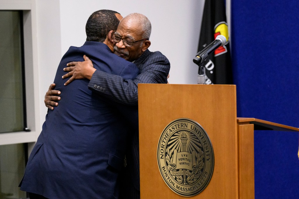 Two men in suits share a warm hug beside a Northeastern University podium during a celebratory event.