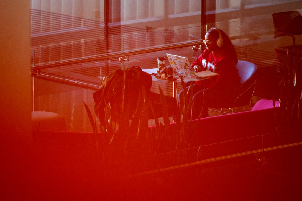 A person wearing headphones studies at a desk in a modern building, surrounded by warm lighting and architectural details.