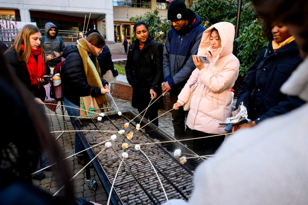 A group of people gathers outdoors, roasting marshmallows over a large grill during a campus event.