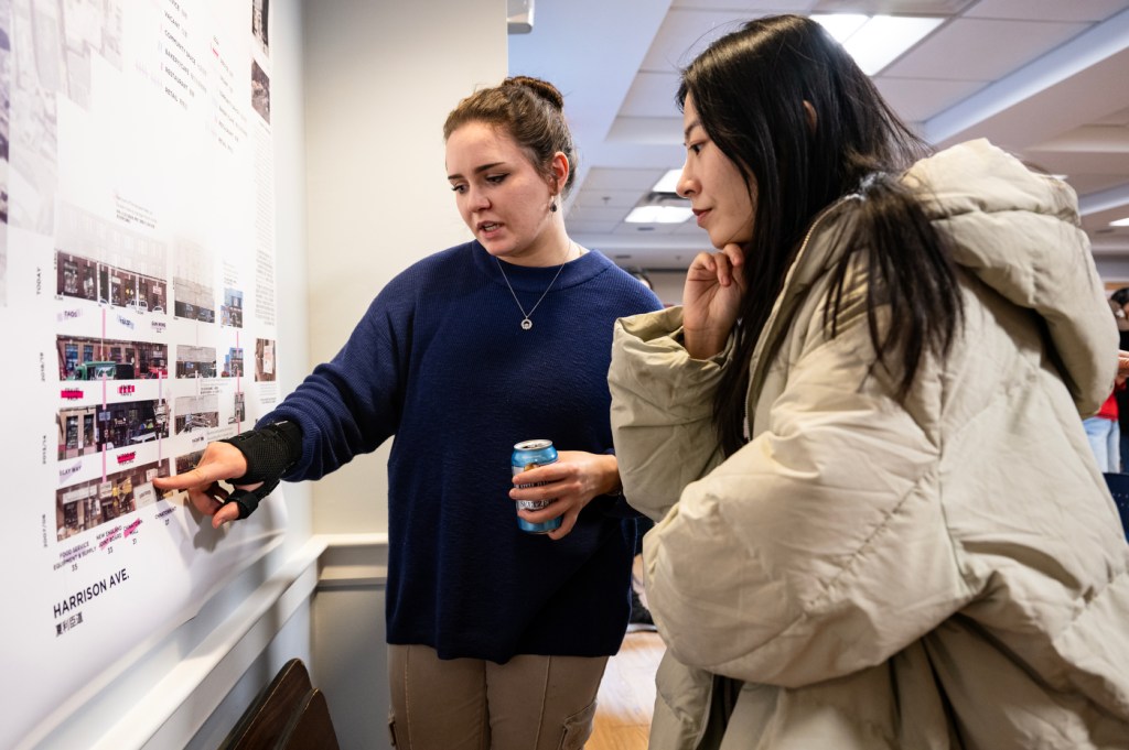 Sasha Hearn wearing a sweater pointing at a project displayed on the wall and explaining it to Feifei Shen, who is leaning in to get a closer look.