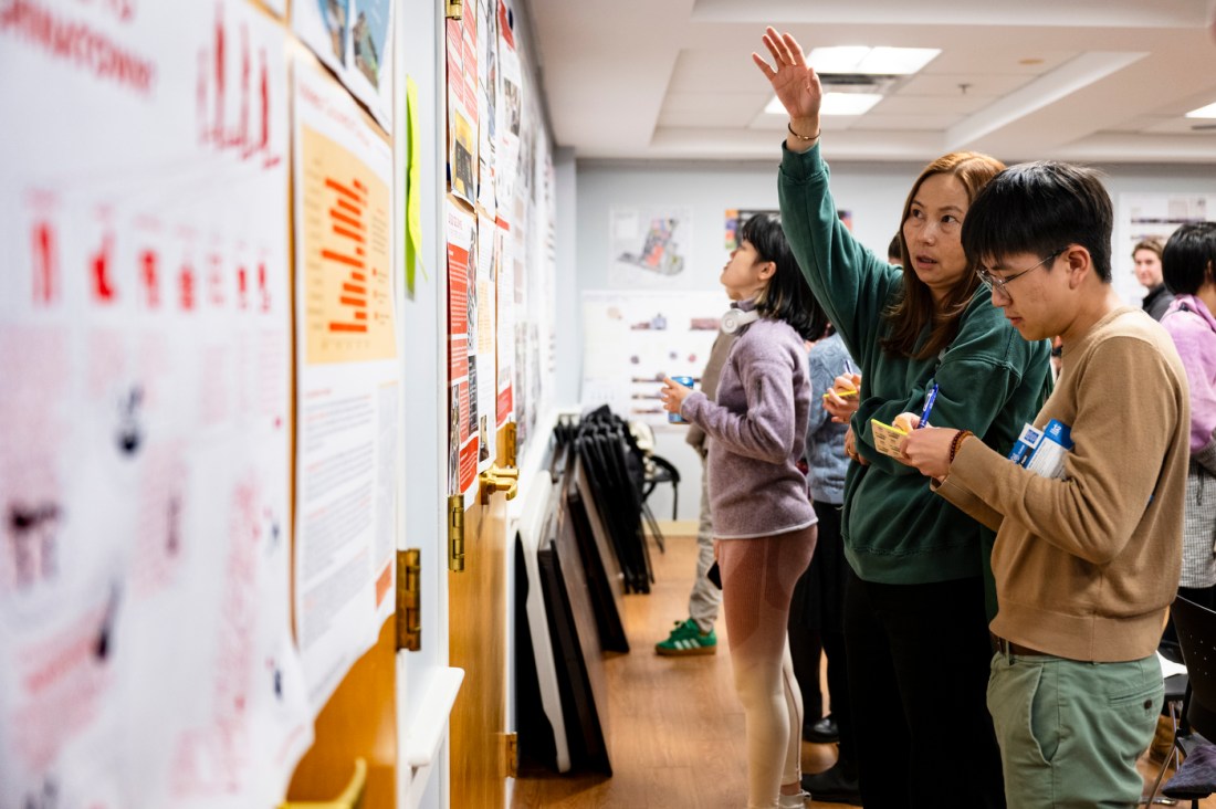 Two students standing in front of a poster displaying architectural projects on it. One of them is gesturing with their right hand.