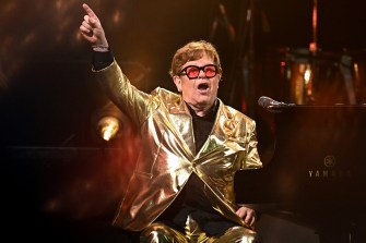 Elton John performing at the piano with a gold suit and pink glasses.