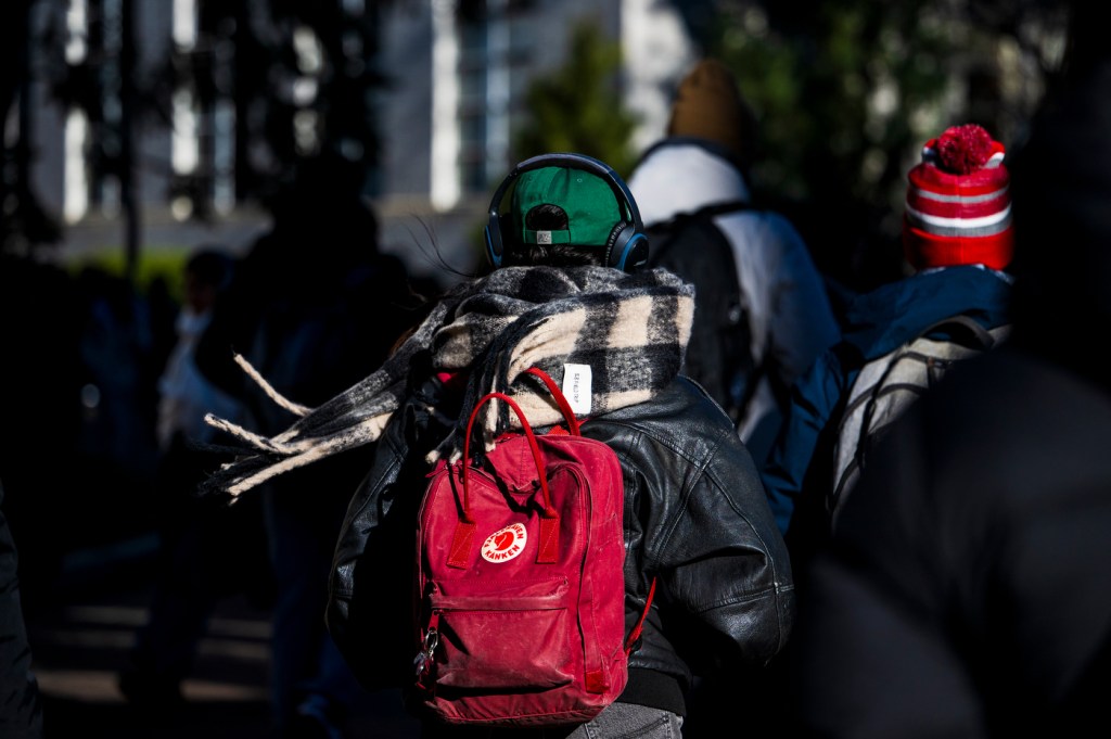 A person wearing a red backpack and a checkered scarf walks outdoors in a crowd on a cold day, surrounded by others dressed warmly in winter clothing.