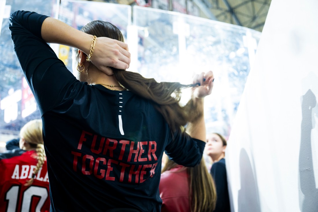 A Northeastern women’s hockey player adjusts her ponytail, wearing a black jacket with "Further Together" written in red on the back.