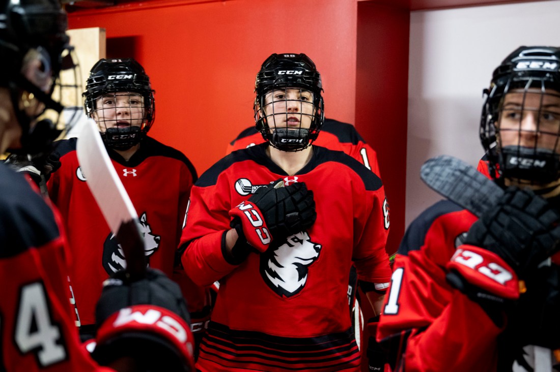 A group of Northeastern women’s hockey players in red jerseys, helmets, and gloves, standing together and preparing to enter the ice.