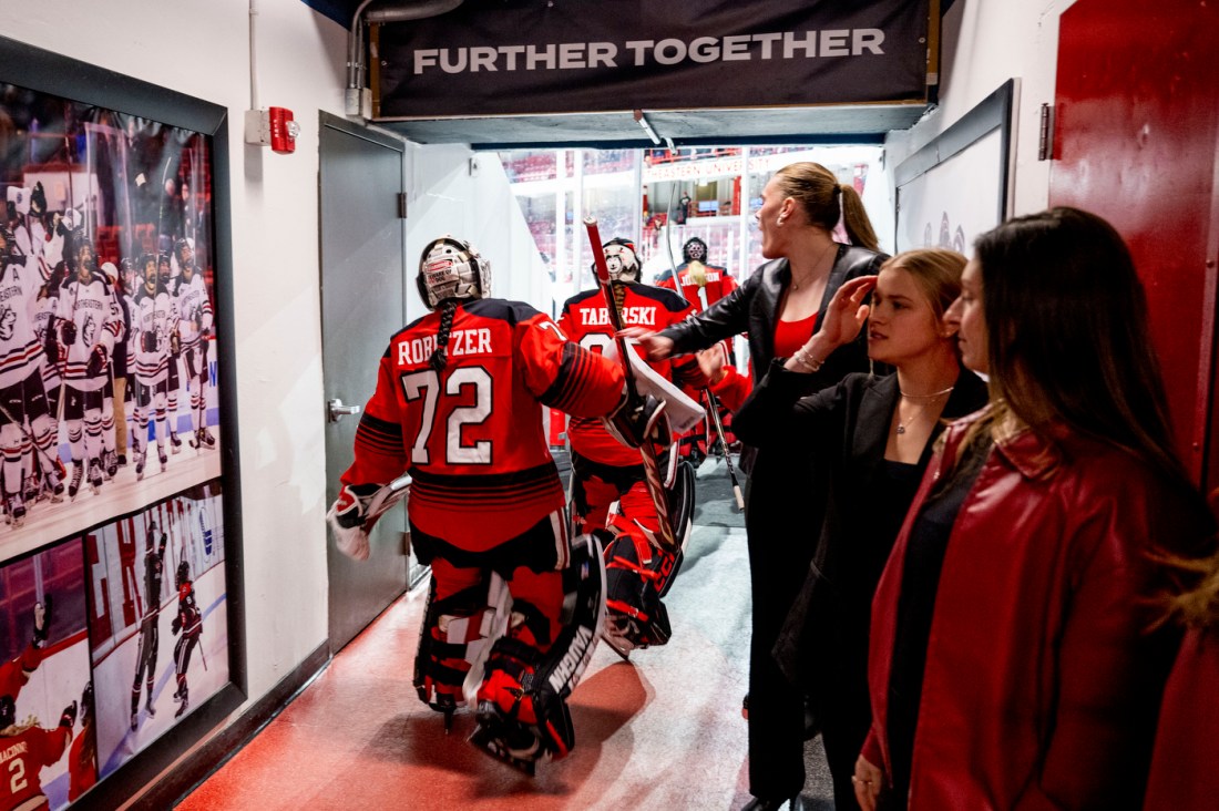 Northeastern women’s hockey goalie and teammates walk under a sign reading "Further Together" as they head onto the ice, with team staff observing nearby.