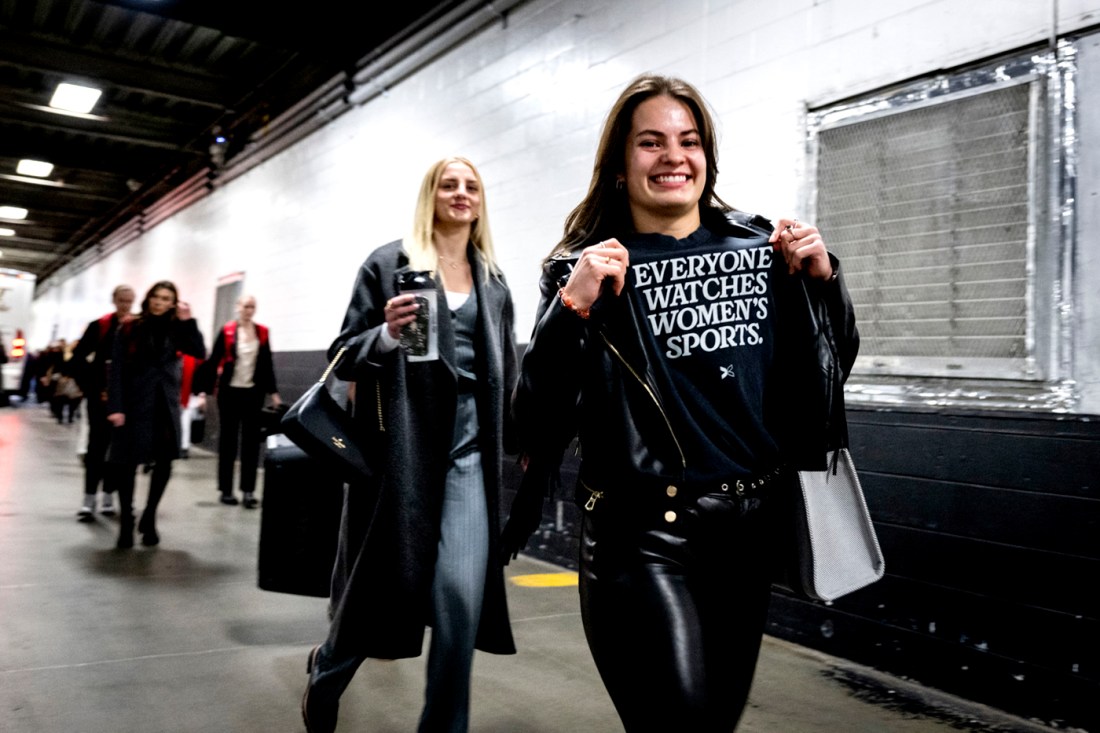 A women's hockey player holding up her black t-shirt that says 'everyone watches women's sports' on it in all caps. 