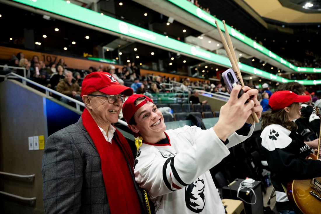 Sam Iannone wearing a backwards red baseball cap and a white hockey jersey taking a selfie with Northeastern's President Aoun who is wearing a red baseball cap and a red scarf.