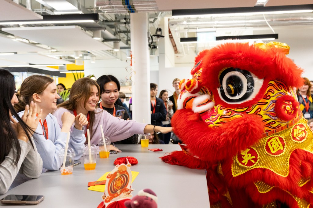 Students gather at a table, smiling and interacting with a vibrant red lion costume during a Lunar New Year celebration on campus.