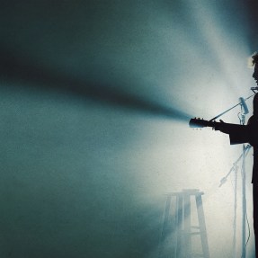 A screen capture from 'A Complete Unknown' of the silhouette of Timothee Chalamet as Bob Dylan playing guitar and singing on stage.