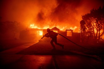 A firefighter carrying a hose in front of a burning home in California.