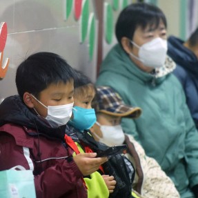 People sit indoors, wearing face masks, with a child holding a device in the foreground.