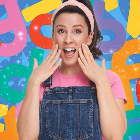 Ms Rachel, in a pink headband, pink t-shirt, and blue overalls, looking at the camera excitedly