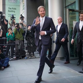 Prince Harry wearing a suit walking out of a building in front of photographers while waving a hand.