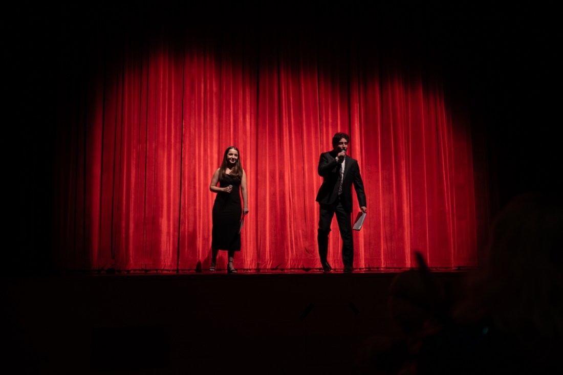 Two students standing on stage next to each other holding microphones in front of a red curtain.