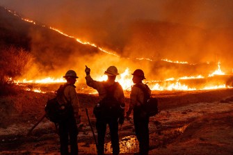 The silhouettes of three fire fighters standing in front of a blazing wildfire in California.