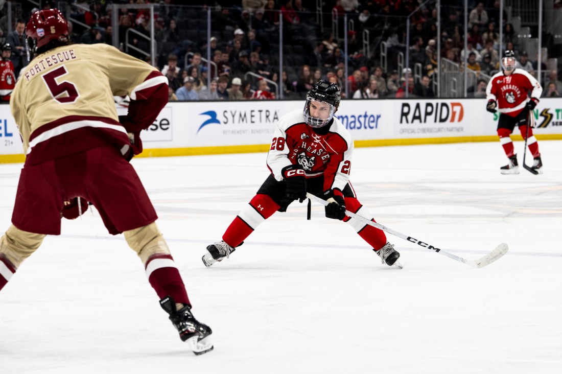 A hockey player in a red and black jersey skating with the puck while being defended by an opposing player in a cream and maroon jersey.