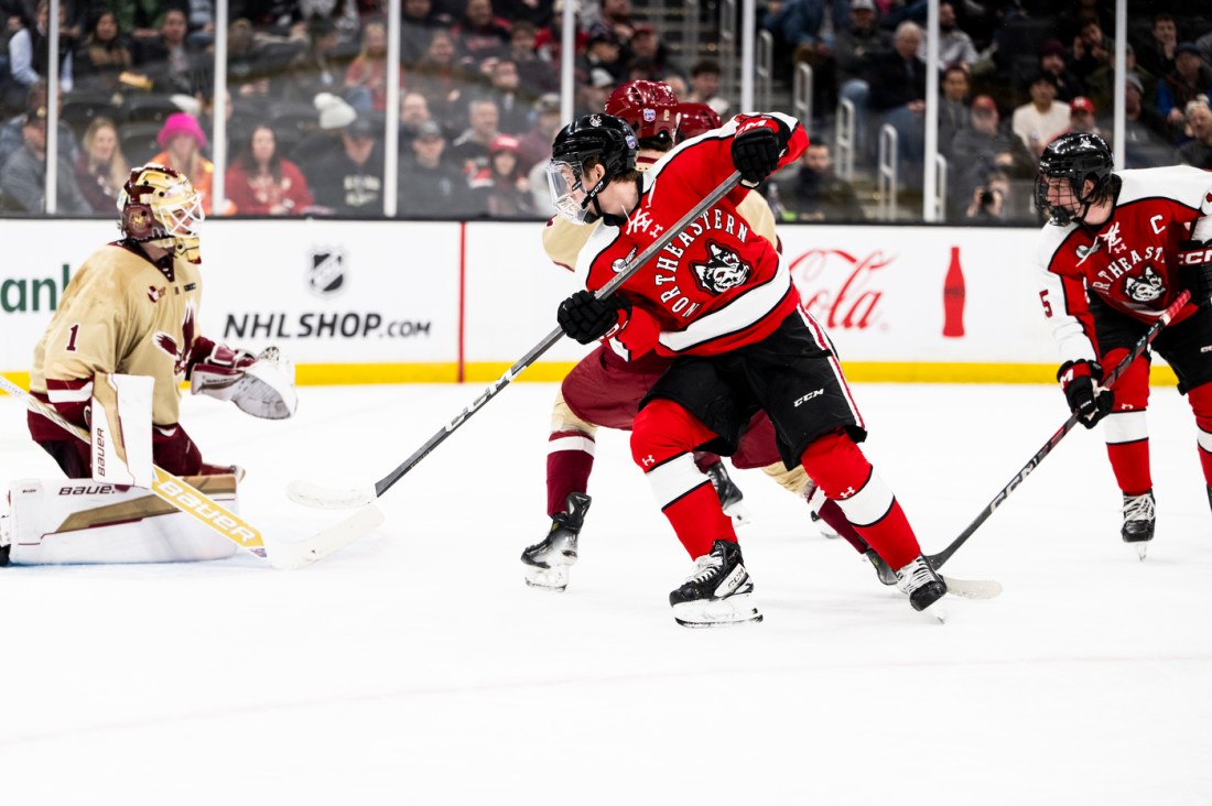 A hockey player in a red and black jersey attempting a shot near the goal, with opposing players and a goalie in cream and maroon jerseys nearby.