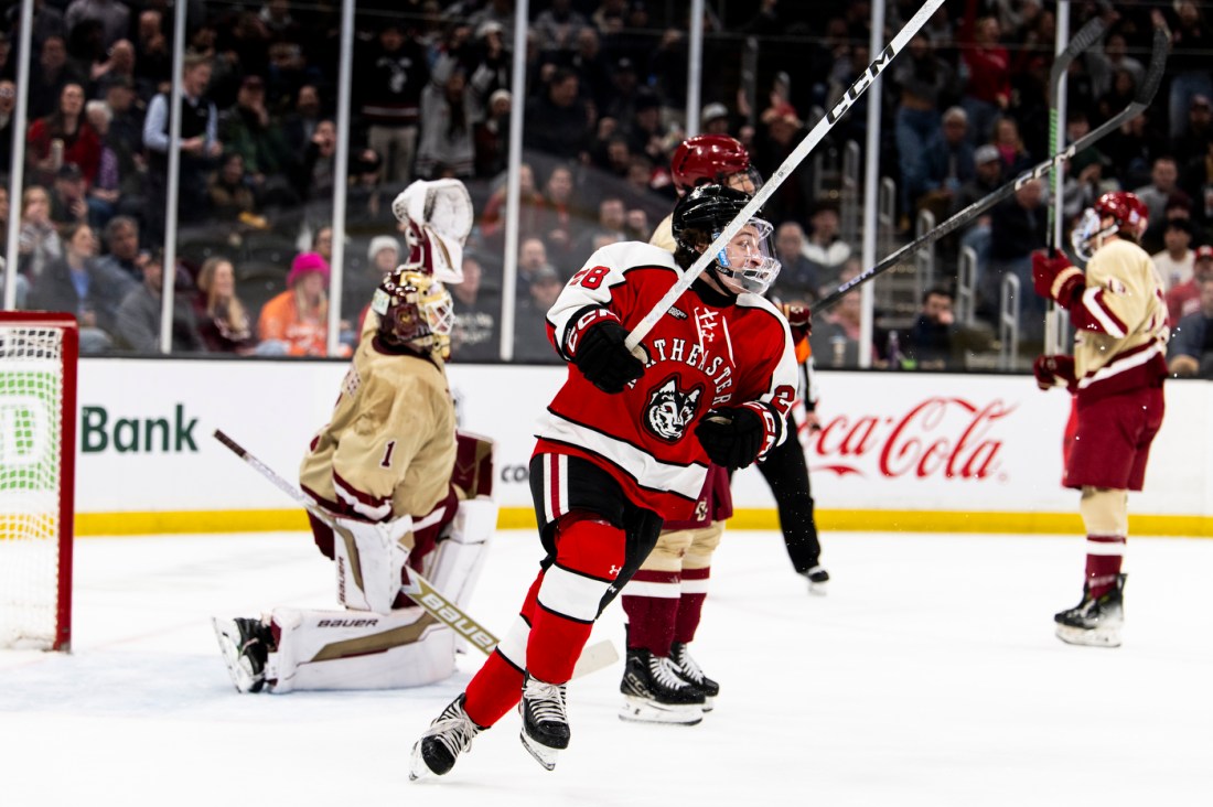 A hockey player in a red and black jersey celebrating on the ice after a goal, with the opposing goalie and other players visible in the background.