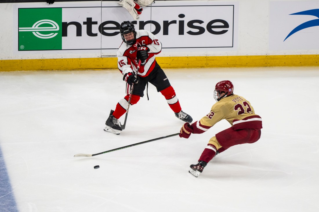 A hockey player in a red and black jersey preparing to take a shot while being defended by an opposing player in cream and maroon.