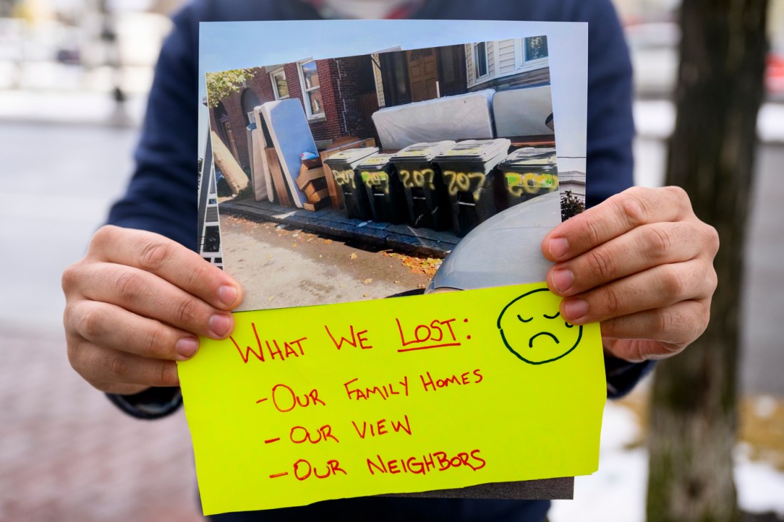 A person holding a printed image of a row of trash outside of a home with a yellow note attached saying 'What We Lost: - Our Family Homes - Our Views - Our neighbors"