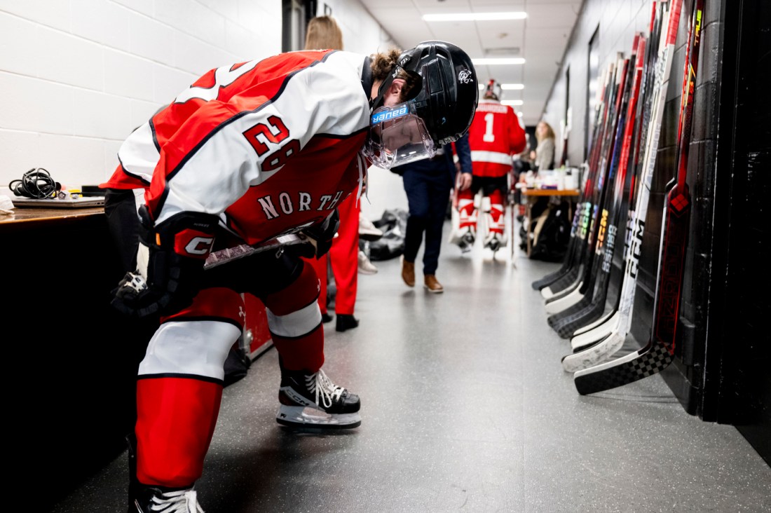 A hockey player in red-and-white gear bends over to adjust their equipment in a hallway lined with hockey sticks, with teammates visible in the background.