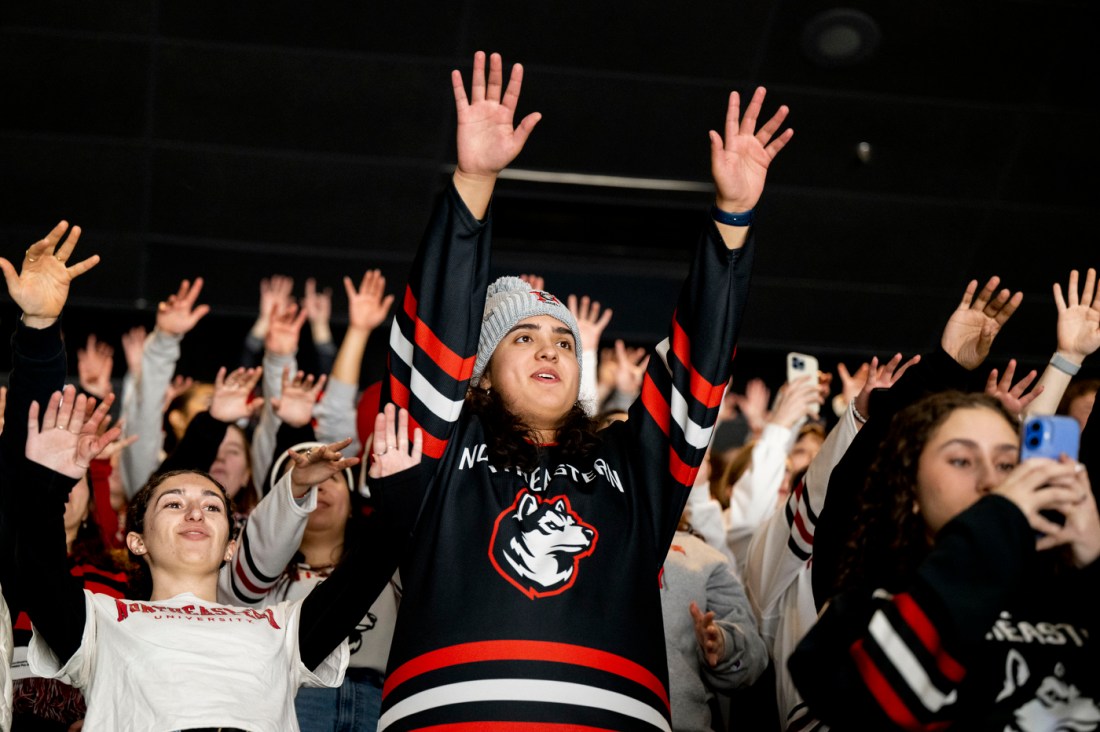 A student wearing a hockey jersey and a grey beanie holds both hands up in the air for a chant.