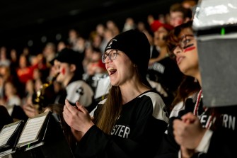 A student wearing a hockey jersey, a black beanie, and glasses claps and cheers in the student section at the men's beanpot semifinal 2025 in TD Garden.