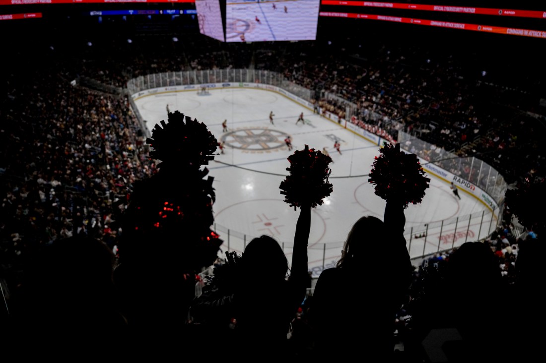 The silhouette of three people holding pom poms cheering while looking down at the rink.