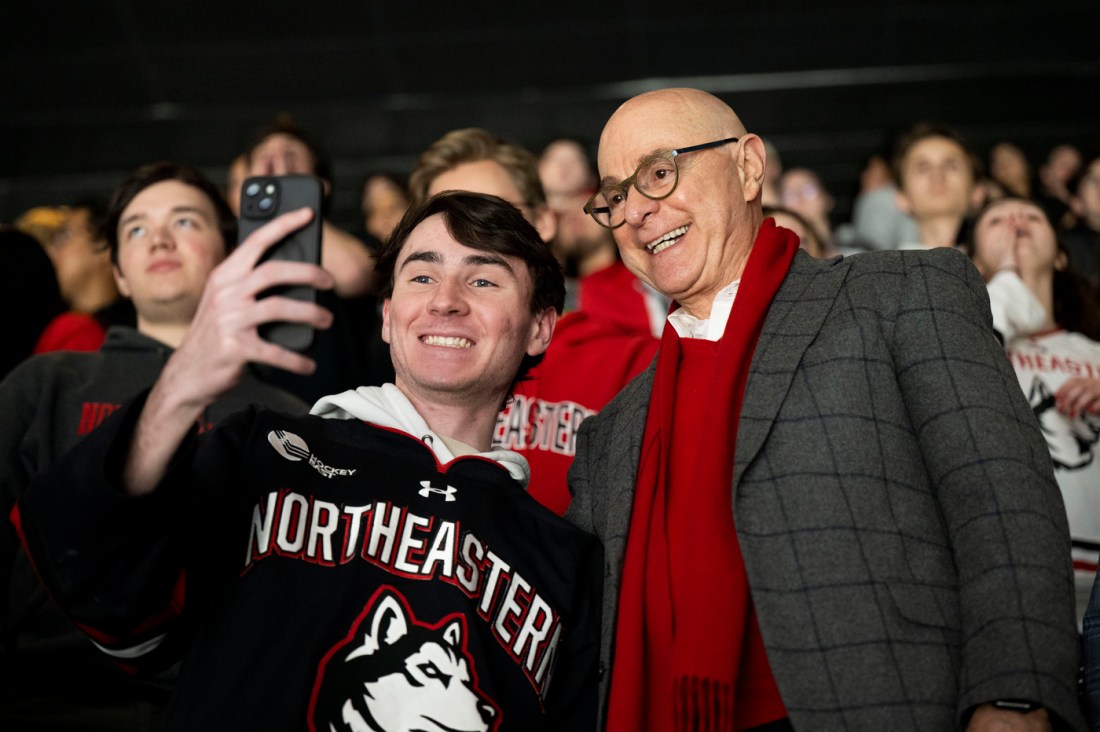 A young person in a Northeastern jersey taking a selfie with an older man in a suit and scarf, both smiling at the camera.